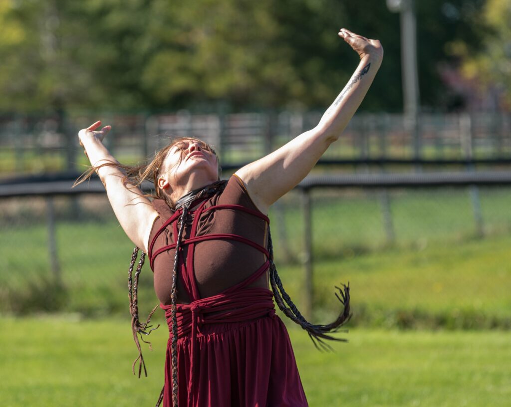 An Indigenous woman performing a solo dance outside on green grass. She has her arms raised, head thrown back.