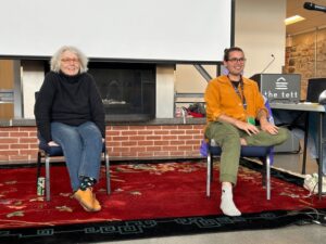 Two people sit on chairs in front of a fireplace with a pull-down screen. One is a woman with white hair, wearing a black top and blue jeans. The other is wearing a burnt orange shirt and green pants