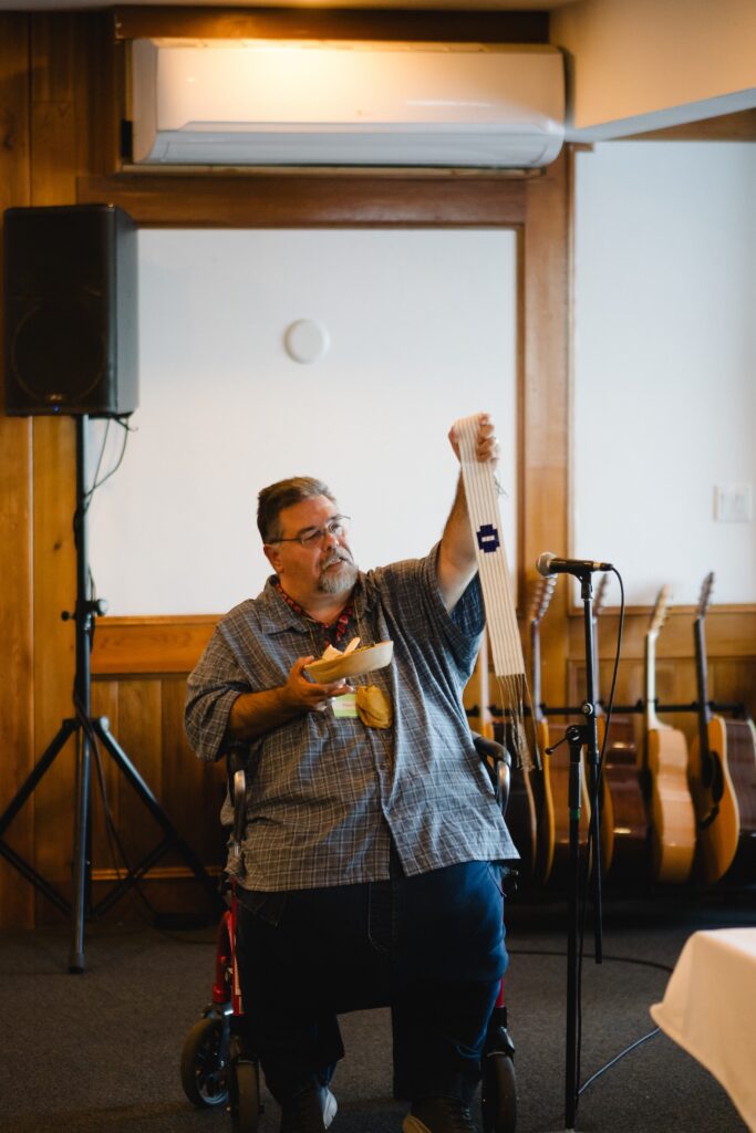 An Indigenous man seated, holding up a bowl and wampum belt, giving a speech.